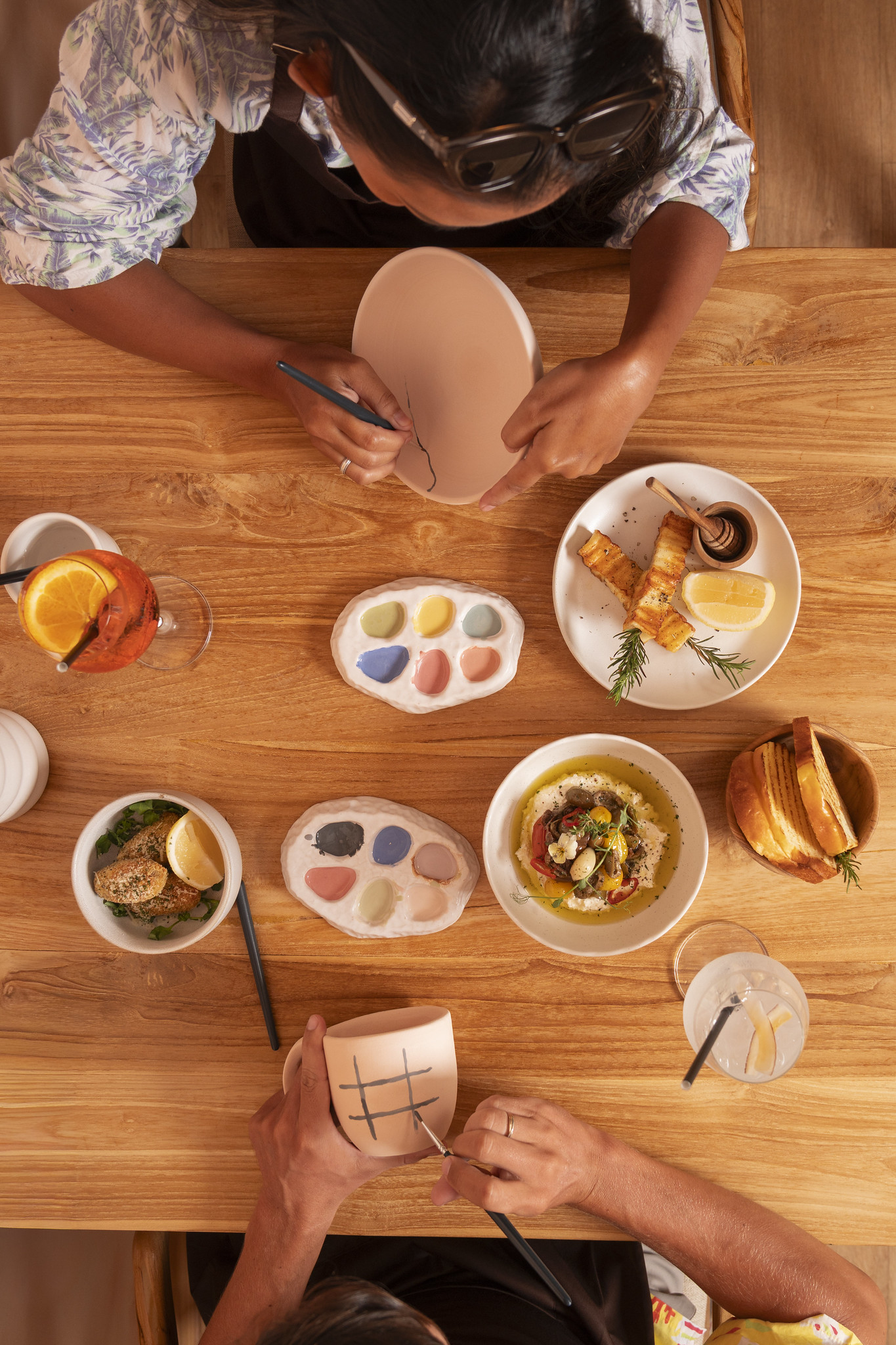 Hands painting ceramic mugs at a wooden table in Klay Bar, with pastel glazes, shared dishes, and drinks creating a relaxed paint-and-sip atmosphere in Canggu.